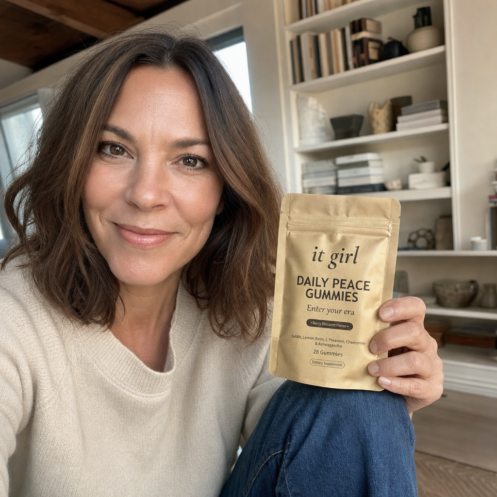 Real photo of a warm professional woman in her late forties with shoulder-length brown hair, a Women's Mental Wellness contributor, sitting in a bright room holding the soft-gold It Girl Daily Peace pouch and smiling at the camera, trusting and calm