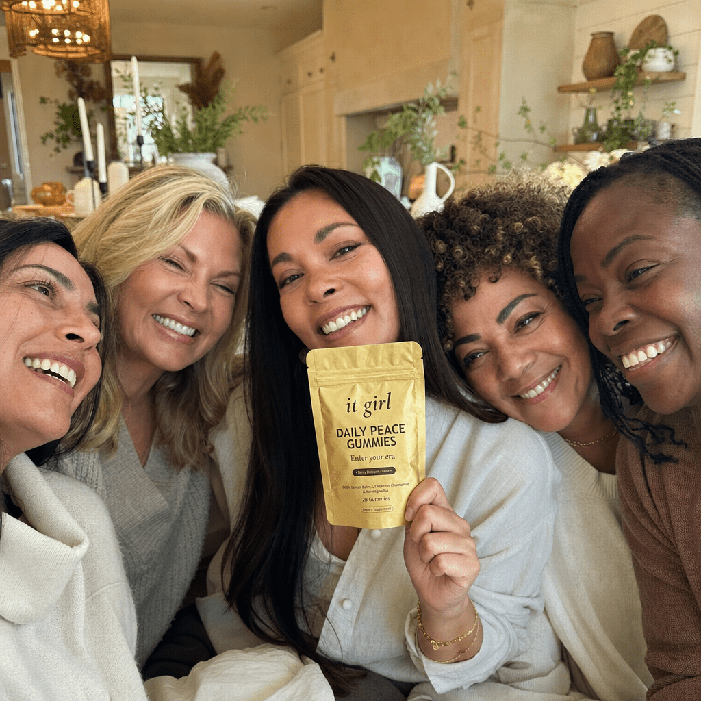 Warm candid group photo of a bunch of real women in their 40s and 50s laughing together in a home setting, one holding up the palm-sized gold It Girl Daily Peace pouch with the front label legible, the women who stopped reaching