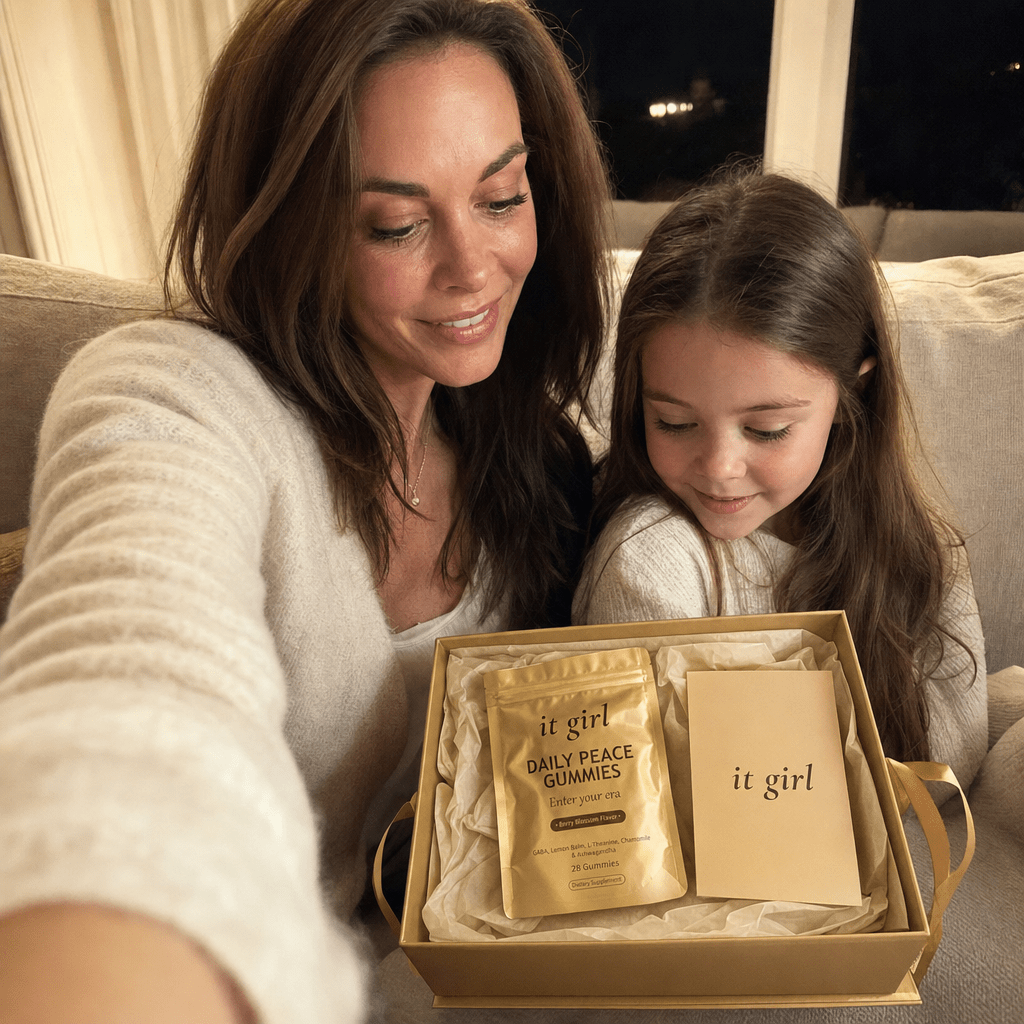 Lauren Hartley taking a nighttime couch selfie with her young daughter as they look at an opened It Girl gift box, the gold Daily Peace pouch nested inside, warm lamp light