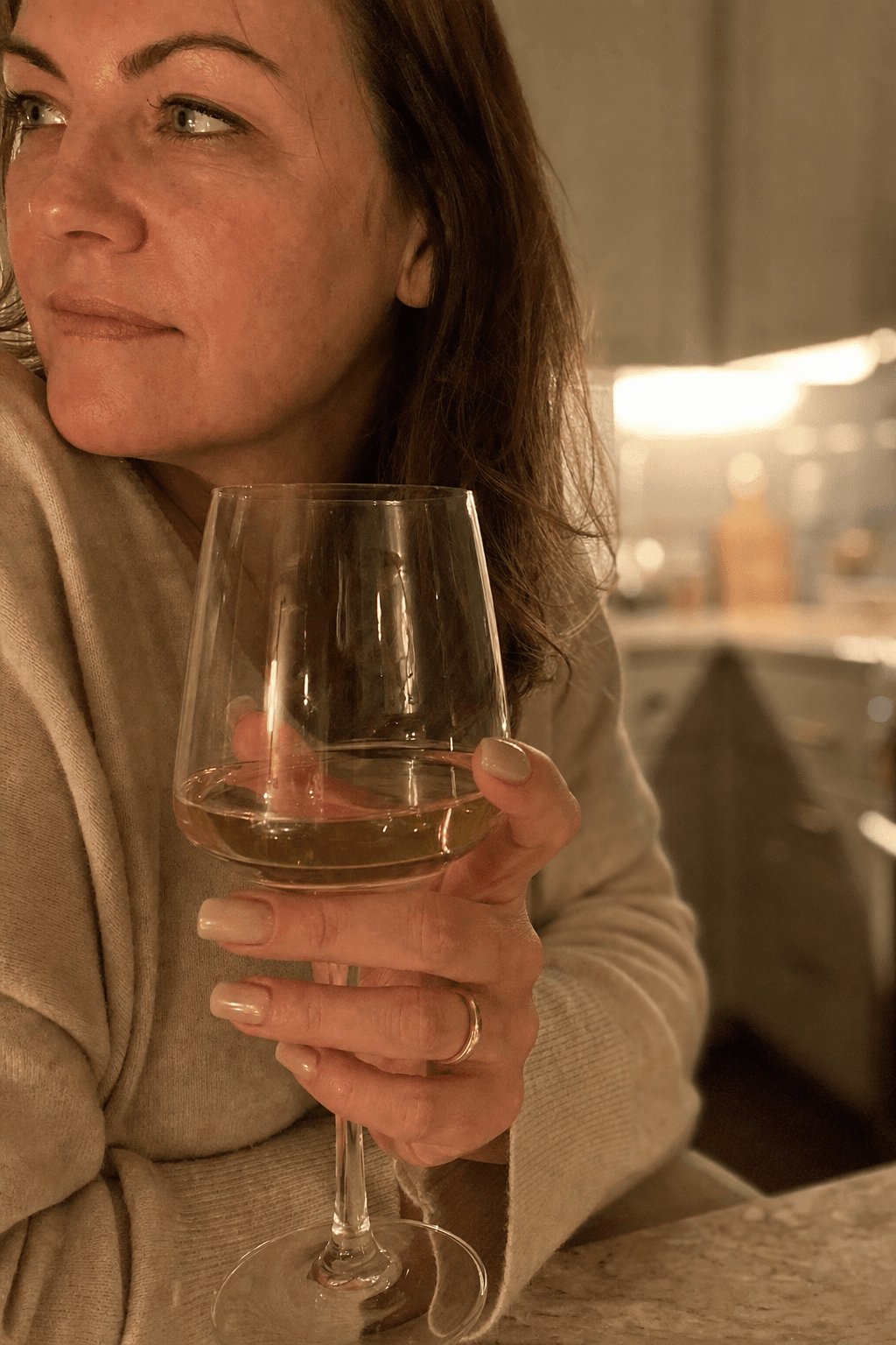Warm macro of a woman's hand wrapped around the stem of a wine glass in gold evening light, the wine glowing, a contemplative and quiet moment, the reach for the calm signal inside the glass
