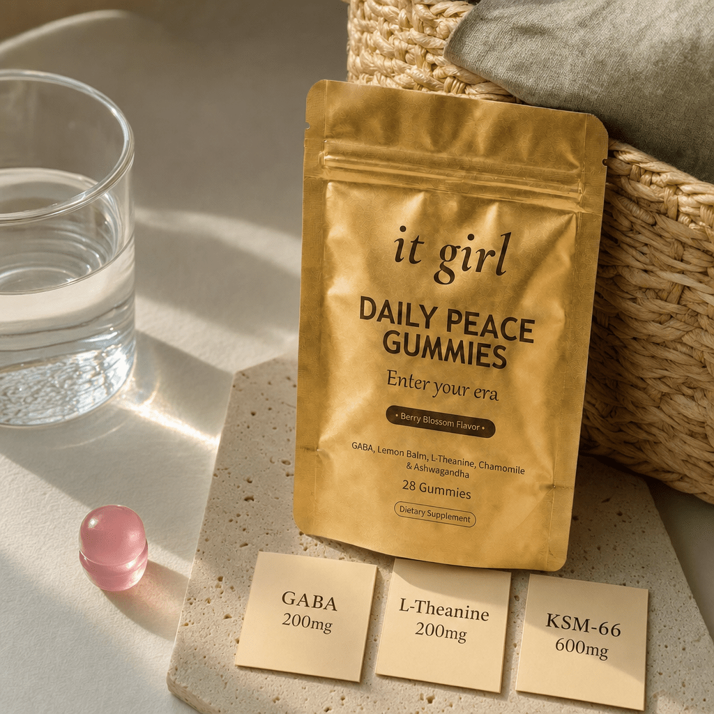 Warm still life of the matte gold It Girl Daily Peace pouch beside a single pink gummy and three dose cards reading GABA 200mg, L-Theanine 200mg and KSM-66 600mg on a cream surface