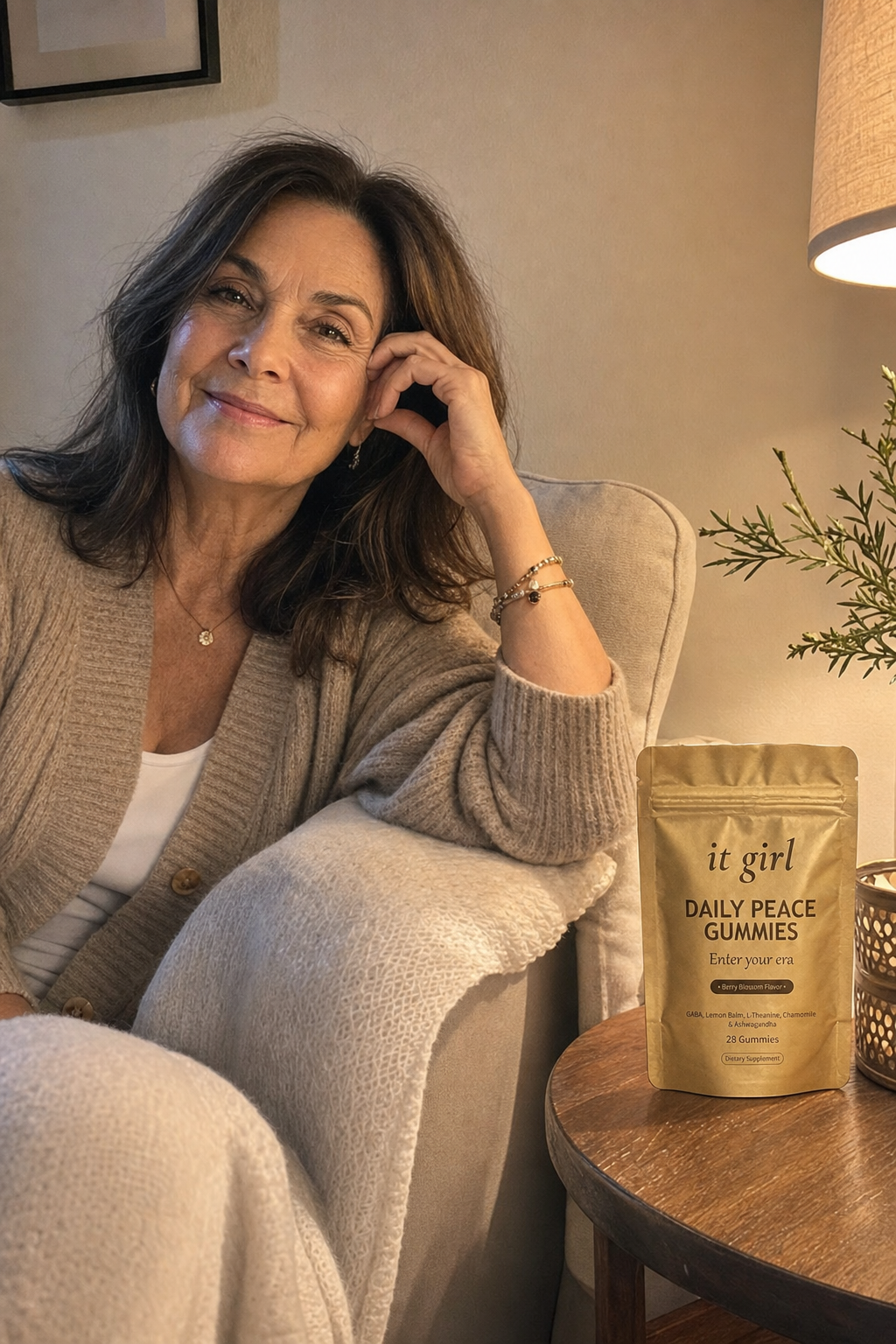 A calm woman in her late fifties at ease on her couch on an ordinary evening, laptop closed, the real matte gold It Girl Daily Peace pouch resting on the side table beside her, the reach finally quiet