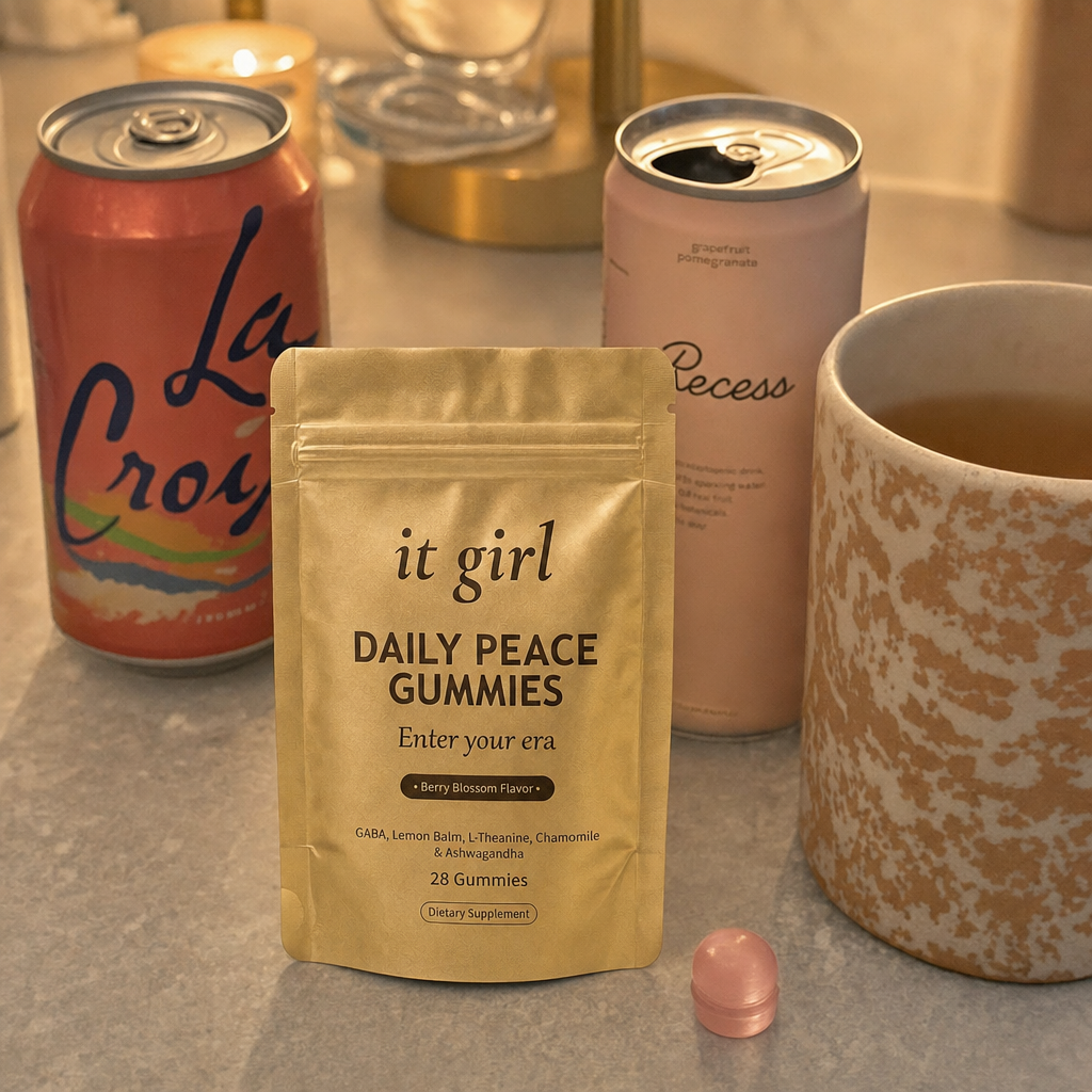 The real matte gold It Girl Daily Peace pouch forward in warm focus on a counter, the abandoned wine swaps, a coral LaCroix can and an adaptogen drink and a mug of cold tea, pushed aside behind it