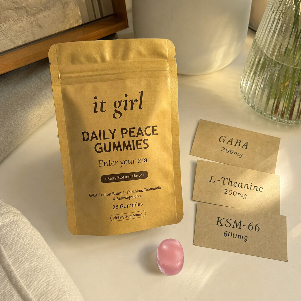 The real matte gold It Girl Daily Peace pouch on a warm cream surface beside a single pink gummy and three named dose cards, GABA 200mg, L-Theanine 200mg, KSM-66 600mg, the studied stack named on the label