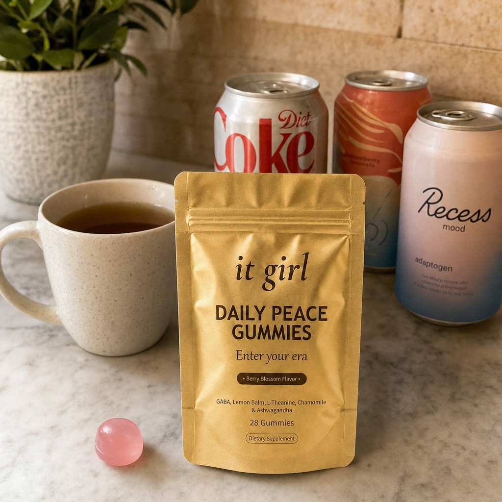 The real matte gold It Girl Daily Peace pouch forward in warm focus on a counter, the abandoned wine swaps, a silver-and-red Diet Coke can and an adaptogen drink and a mug of cold tea, pushed aside behind it