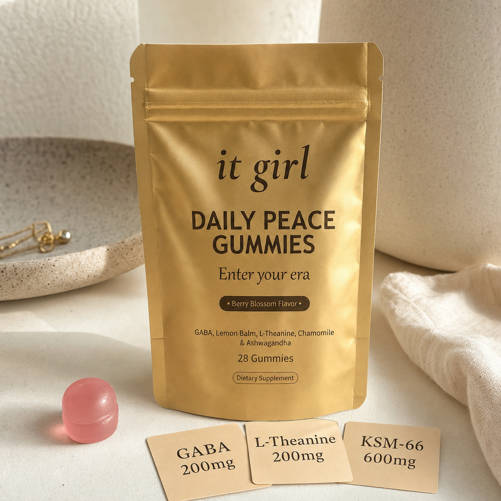 The real matte gold It Girl Daily Peace pouch on a warm cream surface beside a single pink gummy and three named dose cards, GABA 200mg, L-Theanine 200mg, KSM-66 600mg, the studied stack named on the label