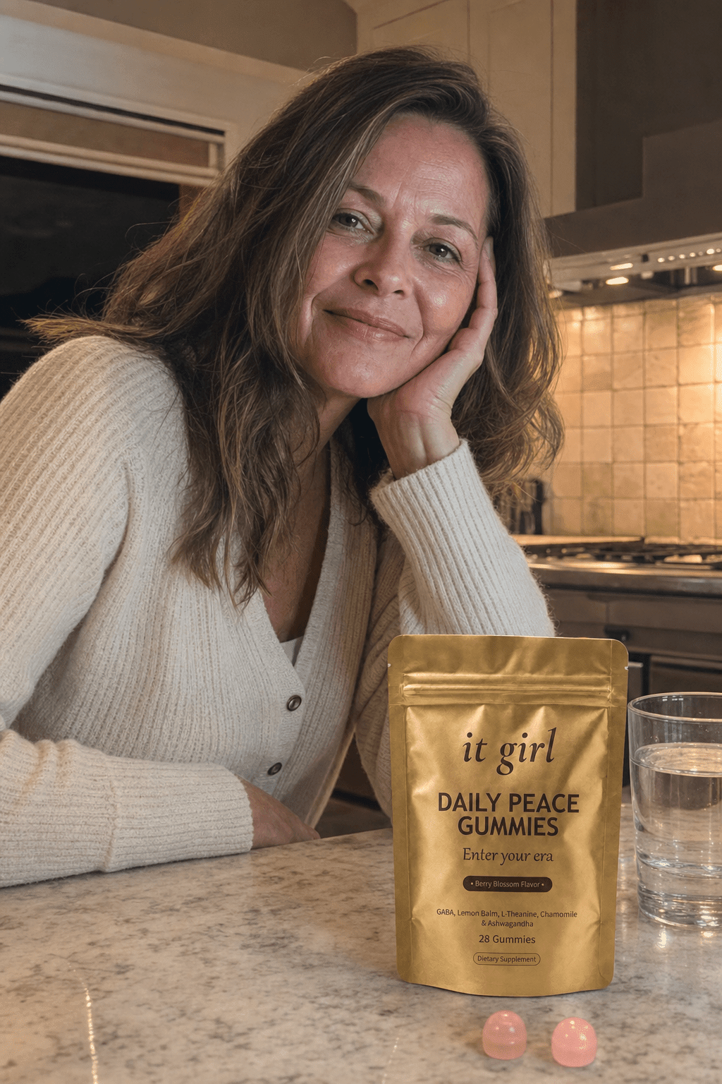 A relaxed woman in her late fifties in a warm evening kitchen, the real gold It Girl Daily Peace pouch on the counter beside her, no wine, calm end of day