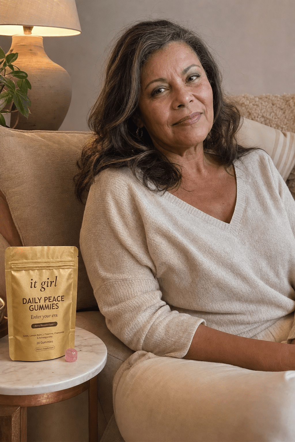 A calm woman in her late fifties at ease on her couch on an ordinary evening, laptop closed, the real matte gold It Girl Daily Peace pouch resting on the side table beside her, the reach finally quiet