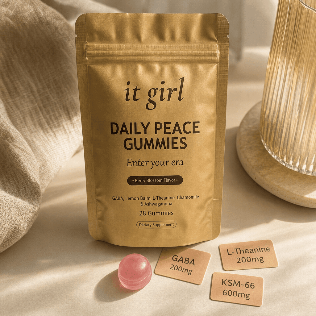 The real matte gold It Girl Daily Peace pouch on a warm cream surface beside a single pink gummy and three named dose cards, GABA 200mg, L-Theanine 200mg, KSM-66 600mg, the studied stack named on the label