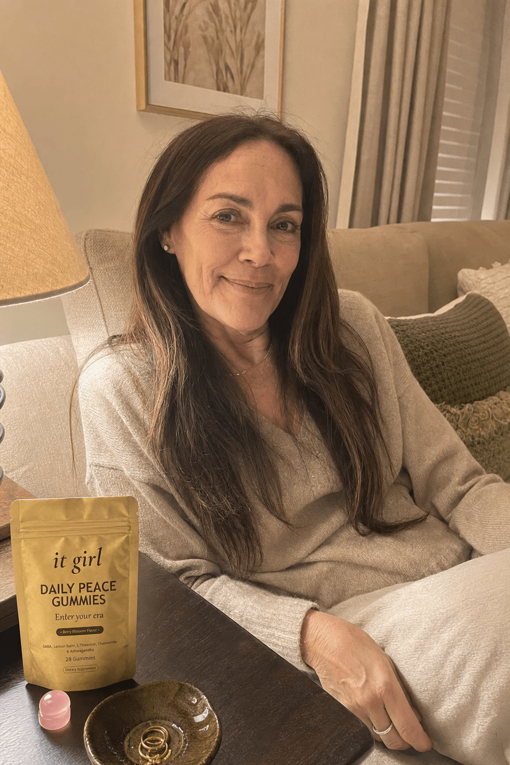 A calm woman in her late fifties at ease on her couch on an ordinary evening, laptop closed, the real matte gold It Girl Daily Peace pouch resting on the side table beside her, the reach finally quiet
