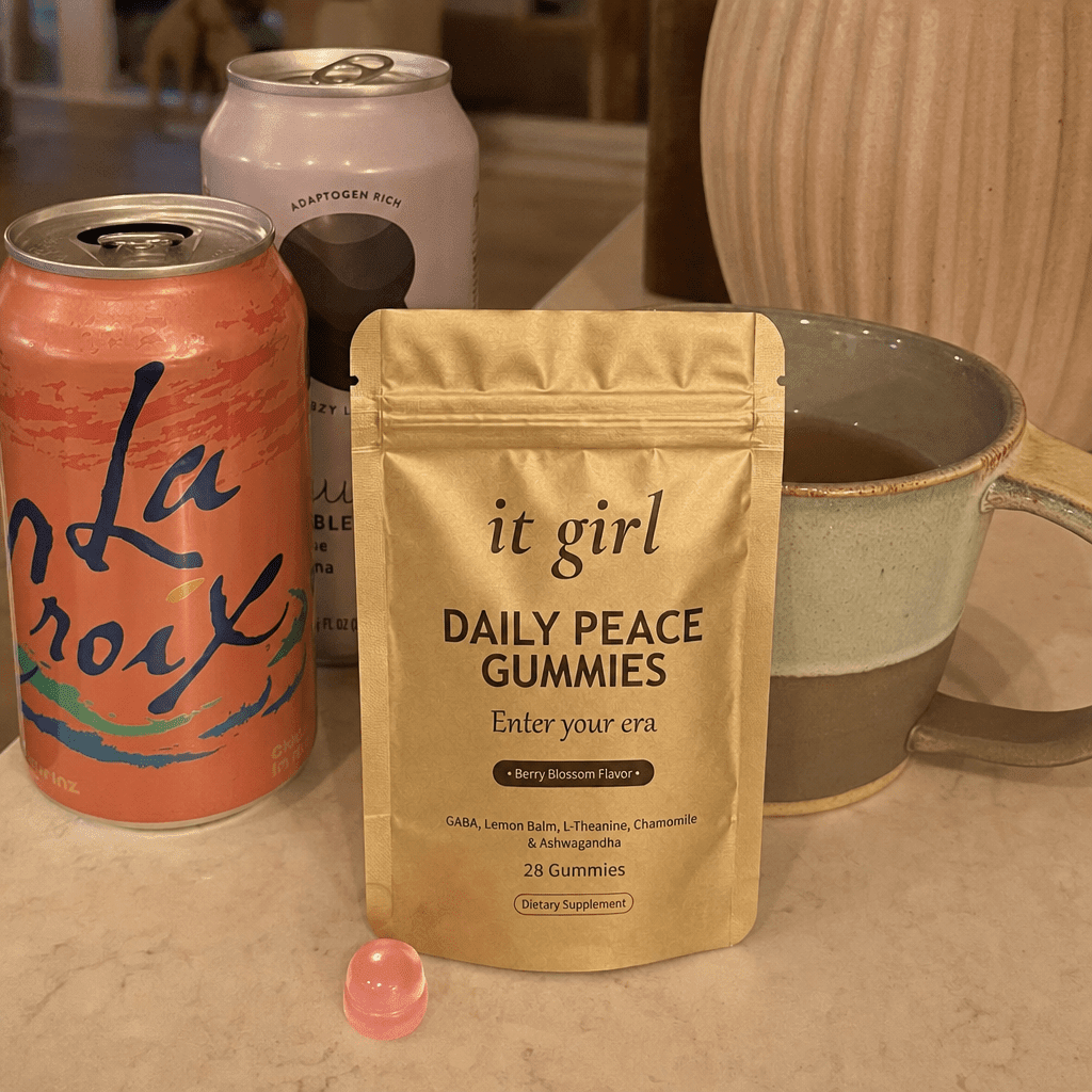 The real matte gold It Girl Daily Peace pouch forward in warm focus on a counter, the abandoned wine swaps, a coral LaCroix can and an adaptogen drink and a mug of cold tea, pushed aside behind it