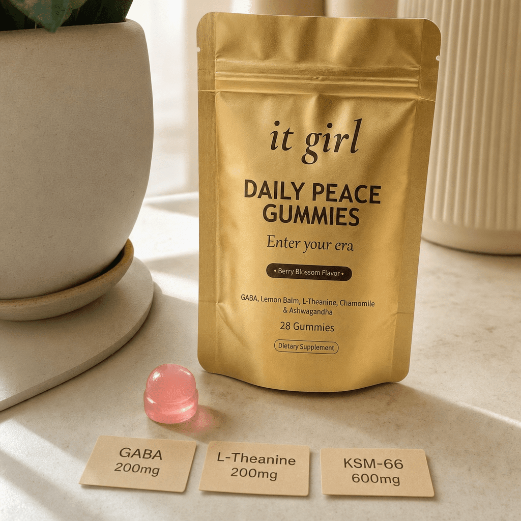 The real matte gold It Girl Daily Peace pouch on a warm cream surface beside a single pink gummy and three named dose cards, GABA 200mg, L-Theanine 200mg, KSM-66 600mg, the studied stack named on the label