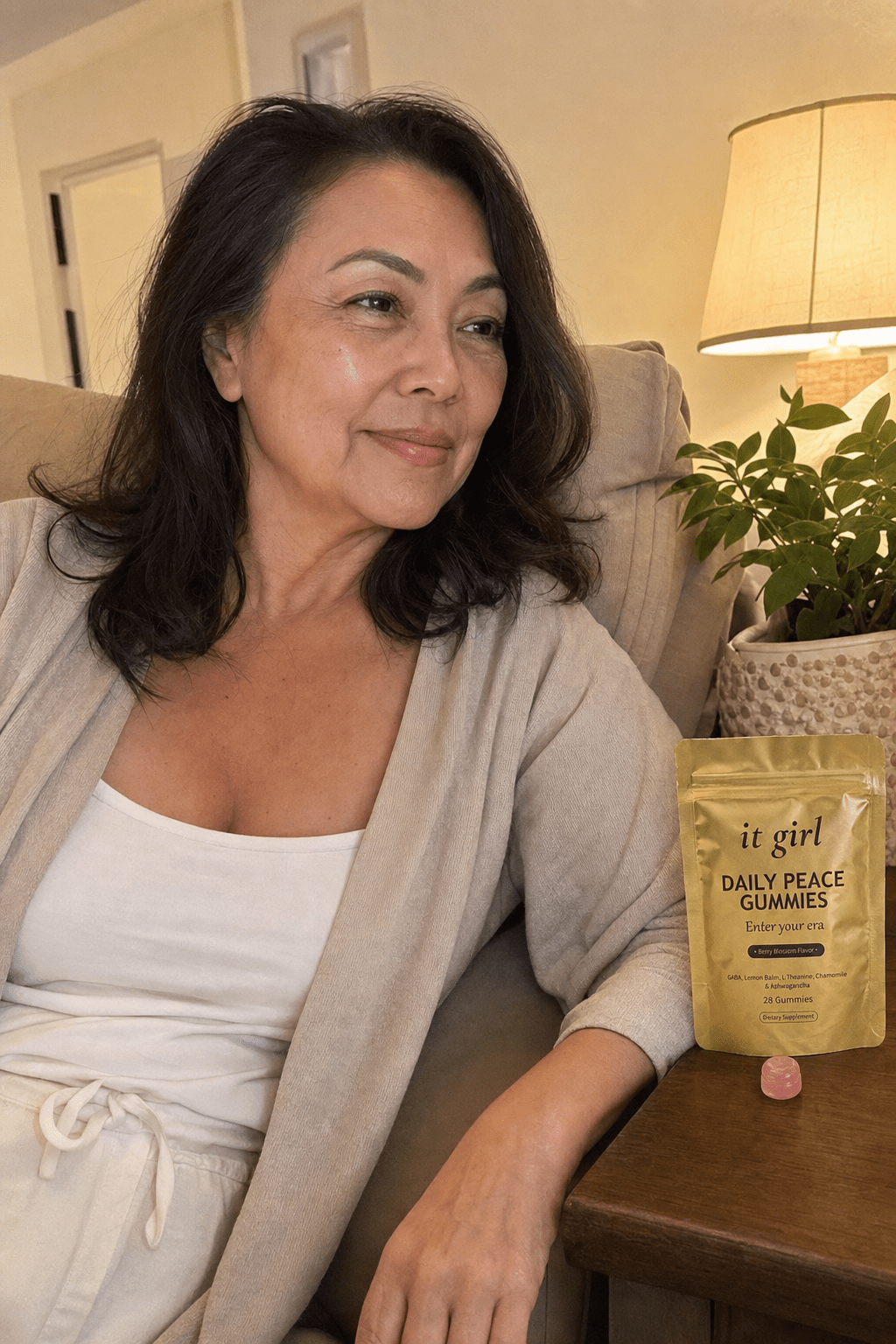 A calm woman in her late fifties at ease on her couch on an ordinary evening, laptop closed, the real matte gold It Girl Daily Peace pouch resting on the side table beside her, the reach finally quiet