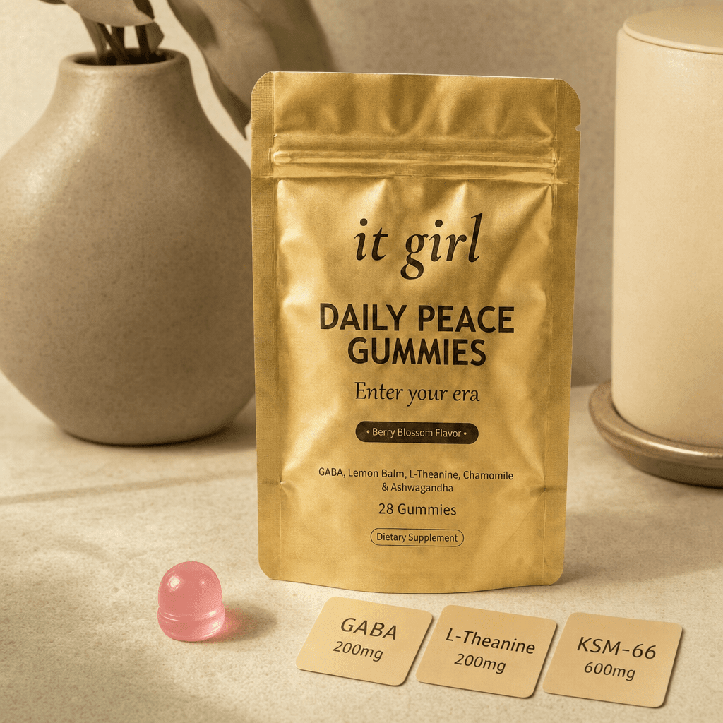 The real matte gold It Girl Daily Peace pouch on a warm cream surface beside a single pink gummy and three named dose cards, GABA 200mg, L-Theanine 200mg, KSM-66 600mg, the studied stack named on the label