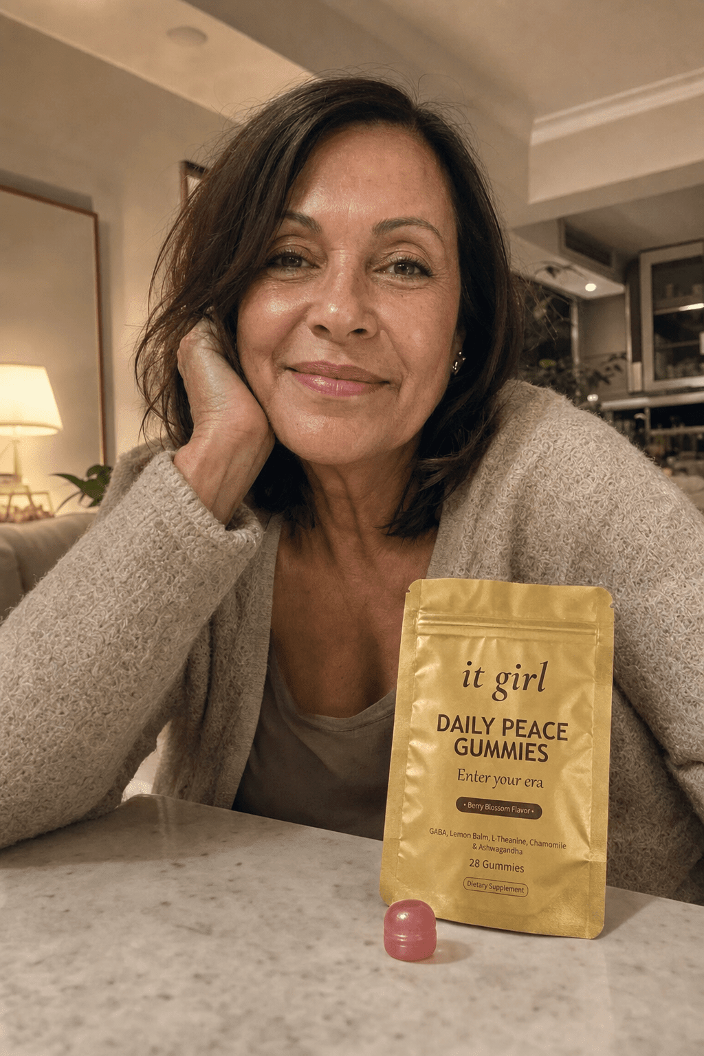 Warm candid evening photo of a relaxed woman in her fifties winding down at home, the real matte-gold It Girl Daily Peace pouch resting on the counter beside her, no wine glass