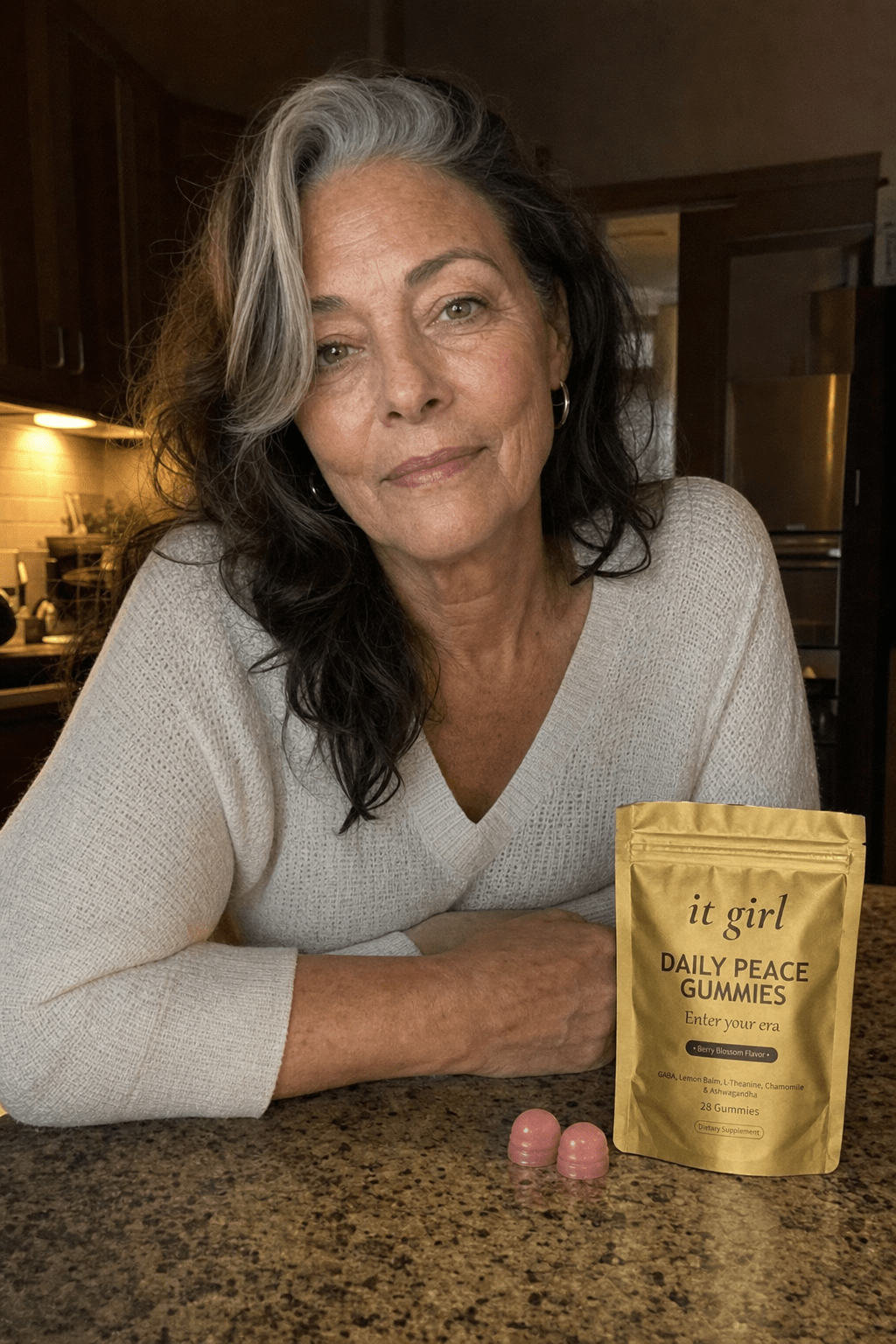Warm evening kitchen photo of a relaxed woman in her late fifties at ease at the counter with the real gold It Girl Daily Peace pouch resting beside her, top rolled open, the calm she was reaching for handed back directly