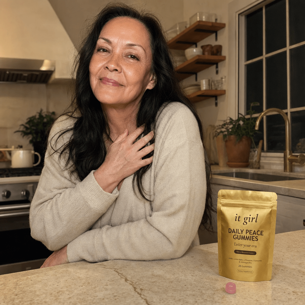 Real woman in her late fifties at ease in a warm-lit evening kitchen, calm and self-forgiving, the gold It Girl Daily Peace pouch resting on the counter, the reach finally understood and no risk in trying