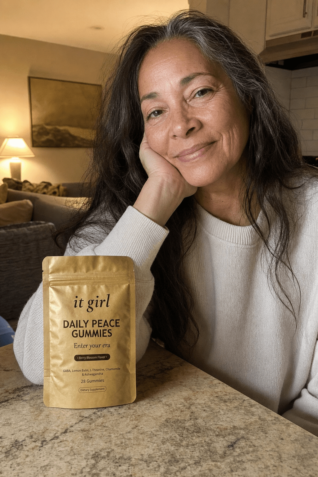 Warm candid evening photo of a relaxed woman in her fifties winding down at home, the real matte-gold It Girl Daily Peace pouch resting on the counter beside her, no wine glass