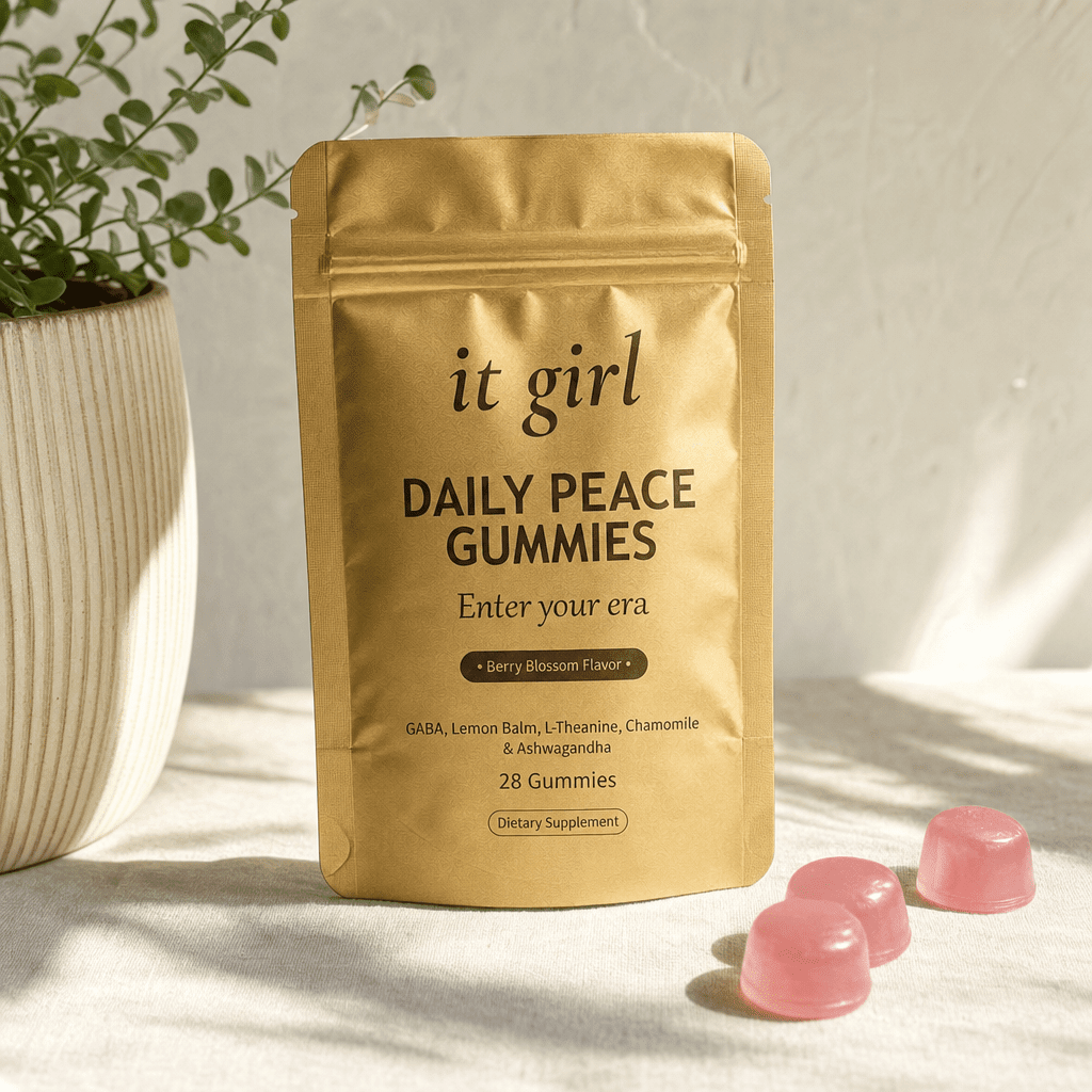 The gold It Girl Daily Peace pouch standing on a cream surface in warm morning light beside its gummies, the studied KSM-66 dose working on cortisol underneath