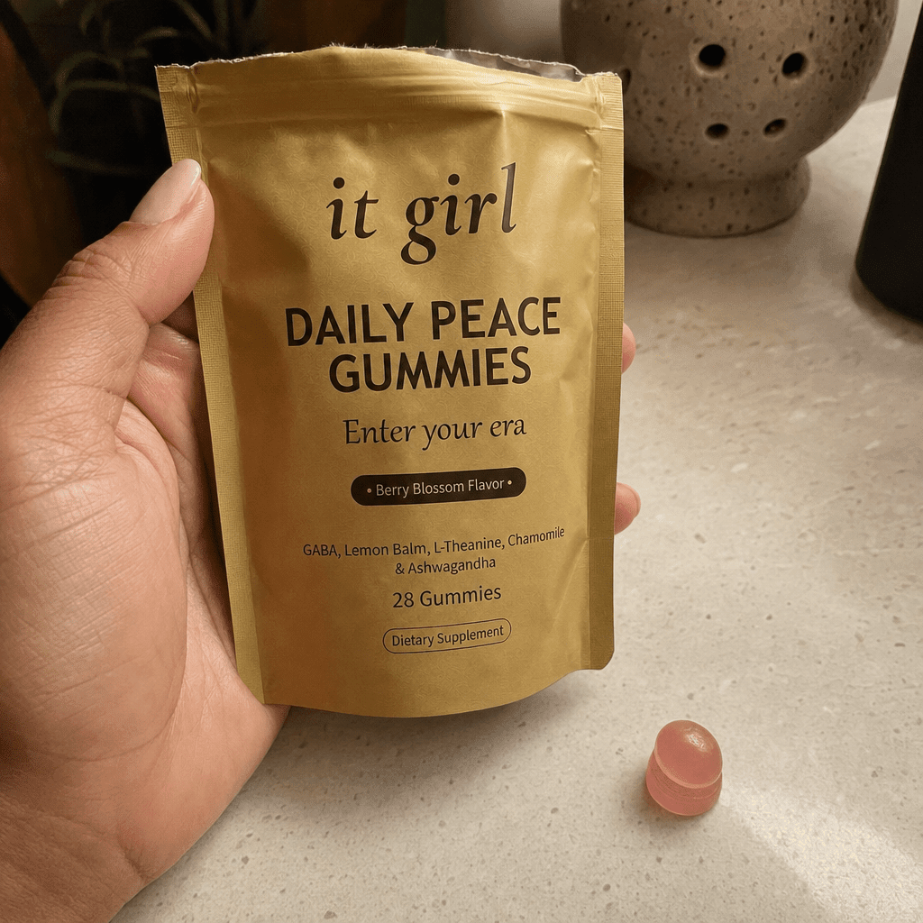 A hand holding the real opened gold It Girl Daily Peace pouch turned to show the legible front label, a pink gummy beside it, the studied doses named on the front