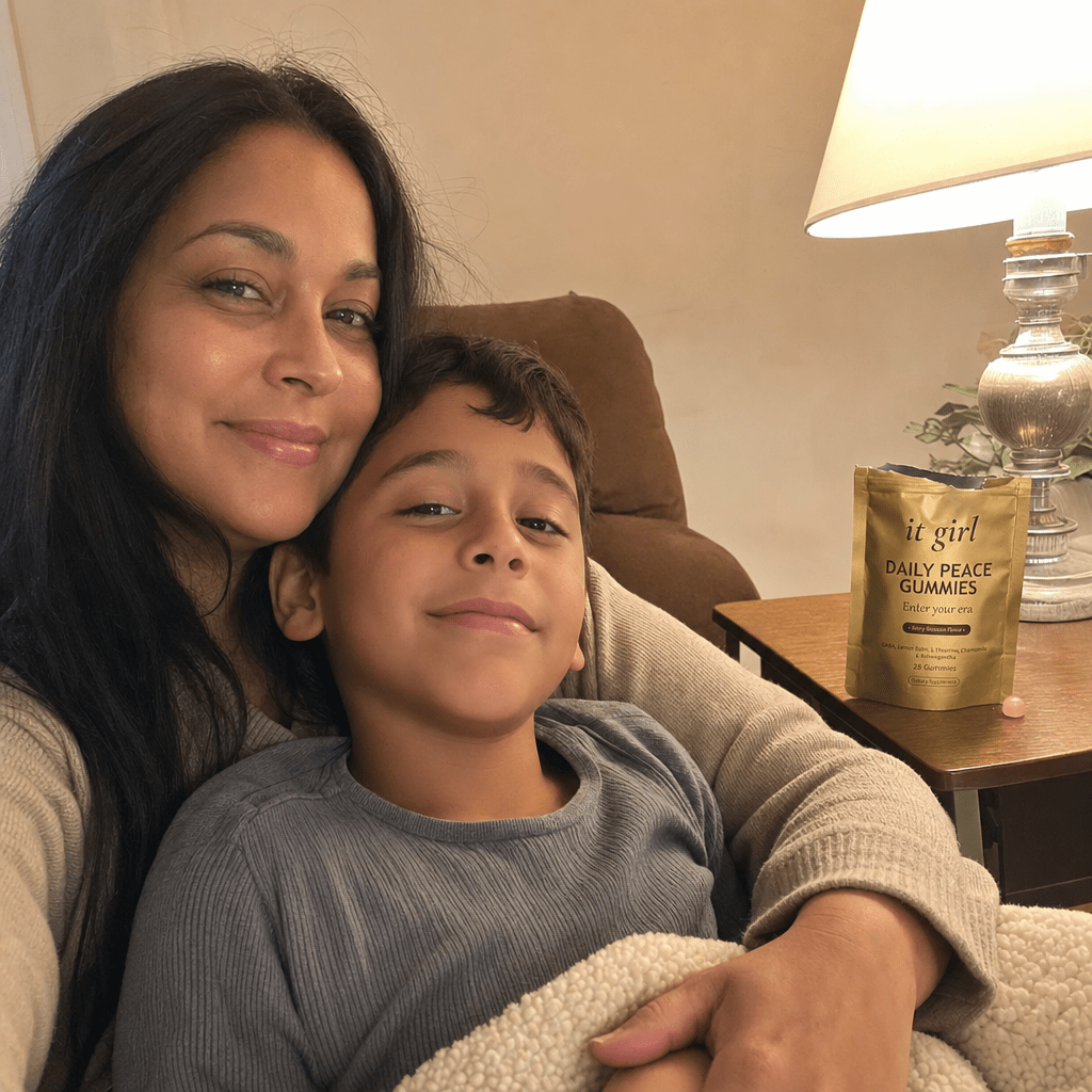 Restored calm mother on the couch in warm evening light with her young son climbed into her lap and staying, the real palm-sized gold It Girl Daily Peace pouch resting on the side table