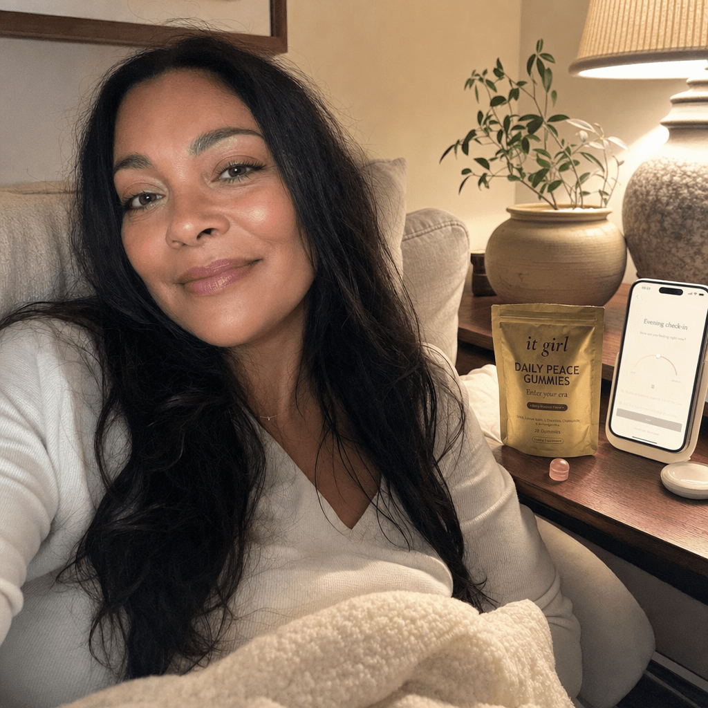 A calm woman settled on her couch in warm lamplight with the gold Daily Peace pouch and a phone showing a quiet evening calm check-in, calm that stays not sedation