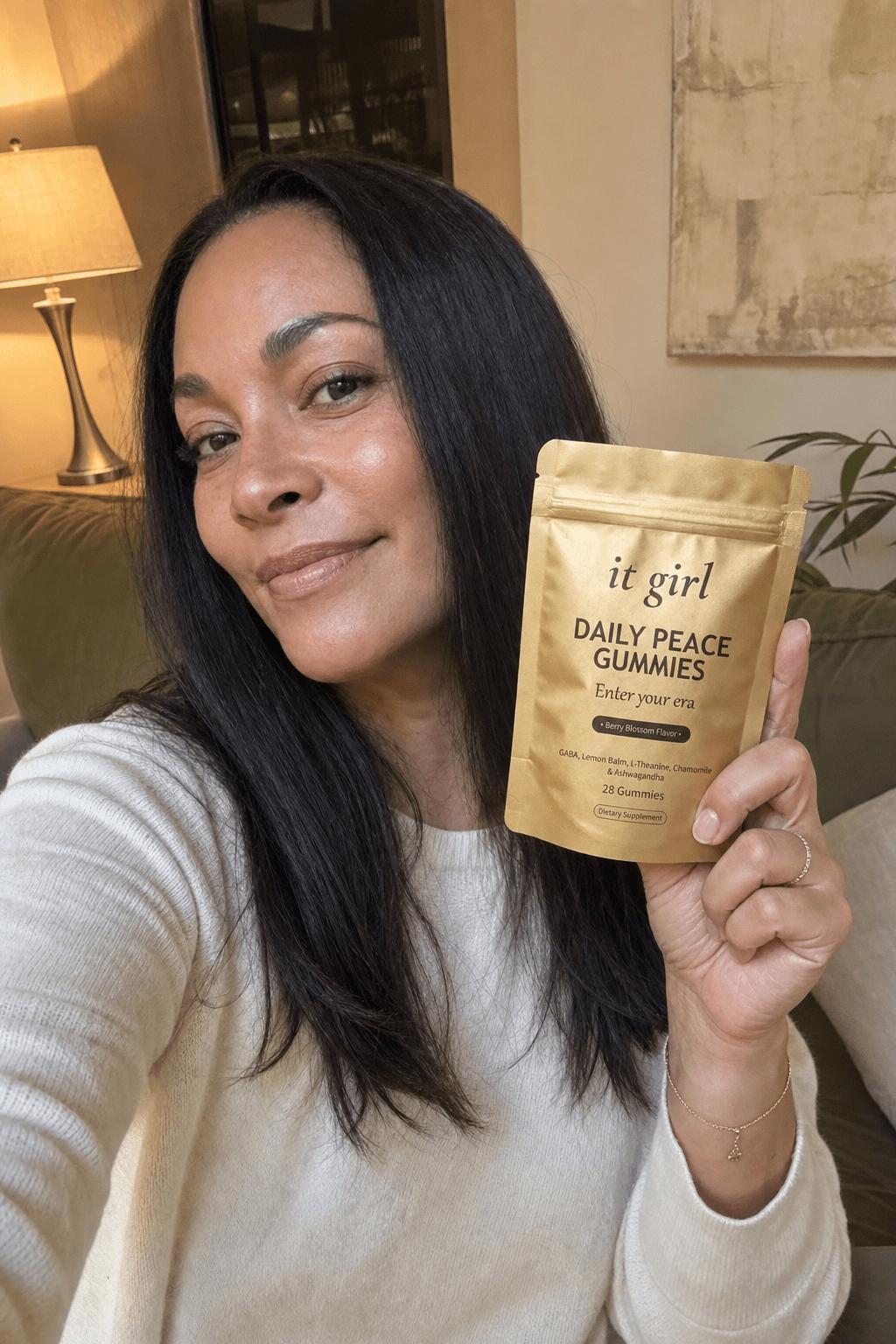 A calm, present woman in her forties in warm early-evening lamplight holding the real gold It Girl Daily Peace pouch with a relieved half-smile, no phone in sight