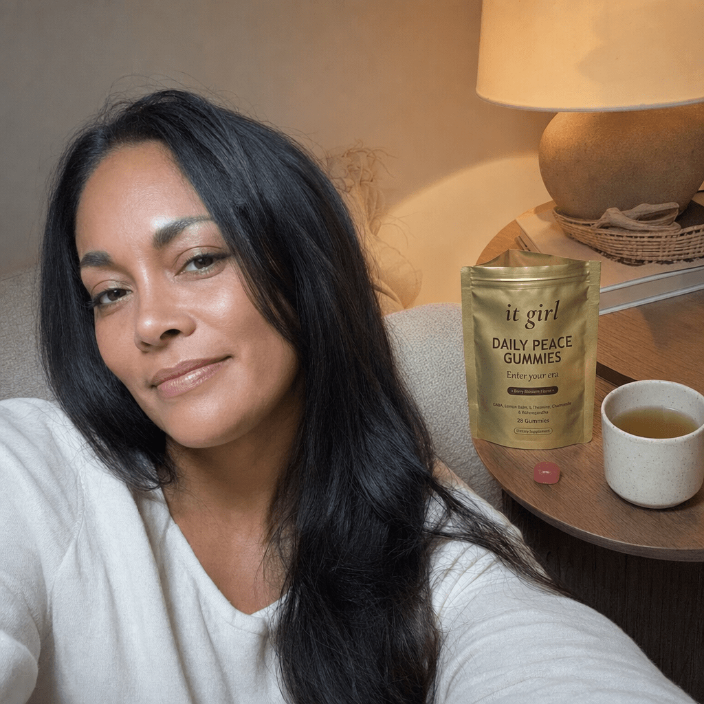 The same calm woman settled on her couch in warm early-evening lamplight with the gold It Girl Daily Peace pouch and a single gummy on the side table, calm not sedation, melatonin-free