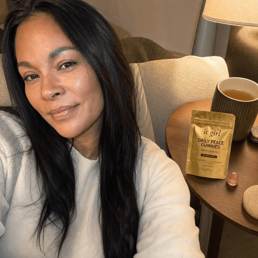 The same calm woman settled on her couch in warm early-evening lamplight with the gold It Girl Daily Peace pouch and a single gummy on the side table, calm not sedation, melatonin-free