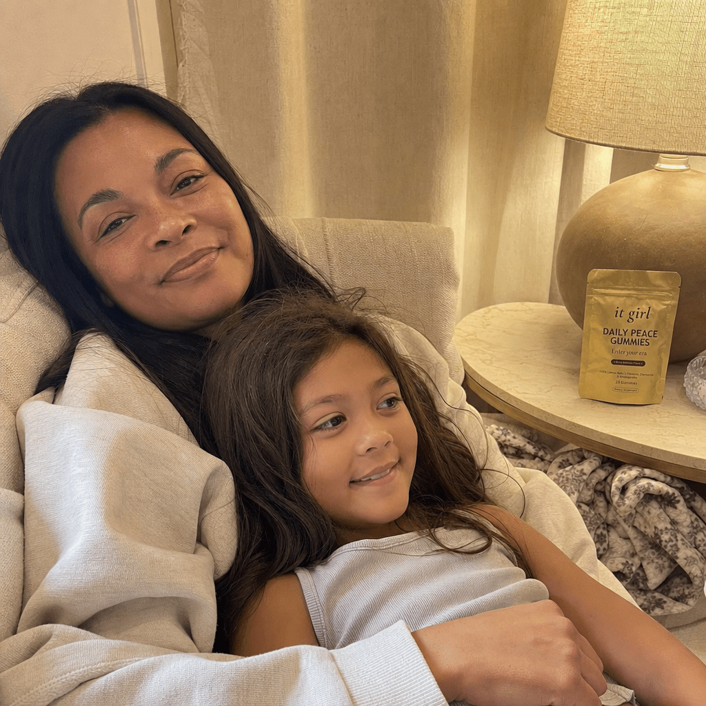 A present mother on the couch in warm lamplight with her young child climbed into her lap and settled in, no bracing, the gold Daily Peace pouch resting in view on the side table