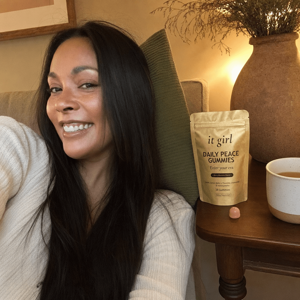 The same calm woman settled on her couch in warm early-evening lamplight with the gold It Girl Daily Peace pouch and a single gummy on the side table, calm not sedation, melatonin-free