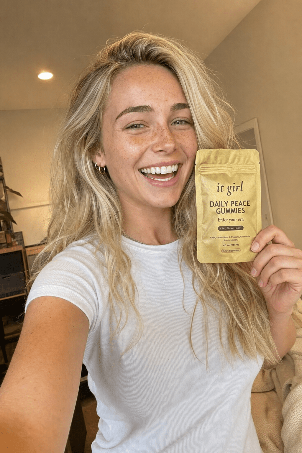 Ultra-realistic candid selfie of Nessa, 24, blonde beach-wave hair and freckles, smiling warmly in soft home light holding the real matte-gold It Girl Daily Peace pouch