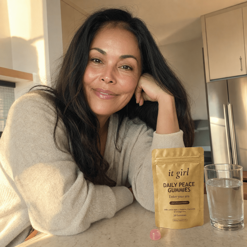 A glowing, restored woman in her early 50s lit from within at a warm kitchen island, her old light back, the gold Daily Peace pouch on the counter and a glass of water, no wine