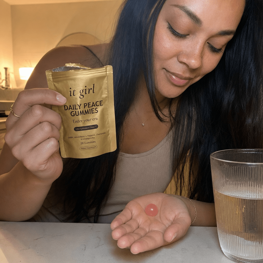 A woman's hand holding the opened matte-gold It Girl Daily Peace pouch tilting a single pink gummy into her palm in warm evening light beside a glass of water, an easy end-of-day ritual