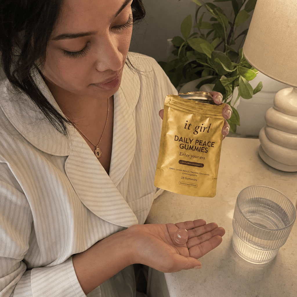 A woman's hand holding the opened matte-gold It Girl Daily Peace pouch with a single pink gummy in warm evening light beside a glass of water on a nightstand, a dollar-a-day evening ritual