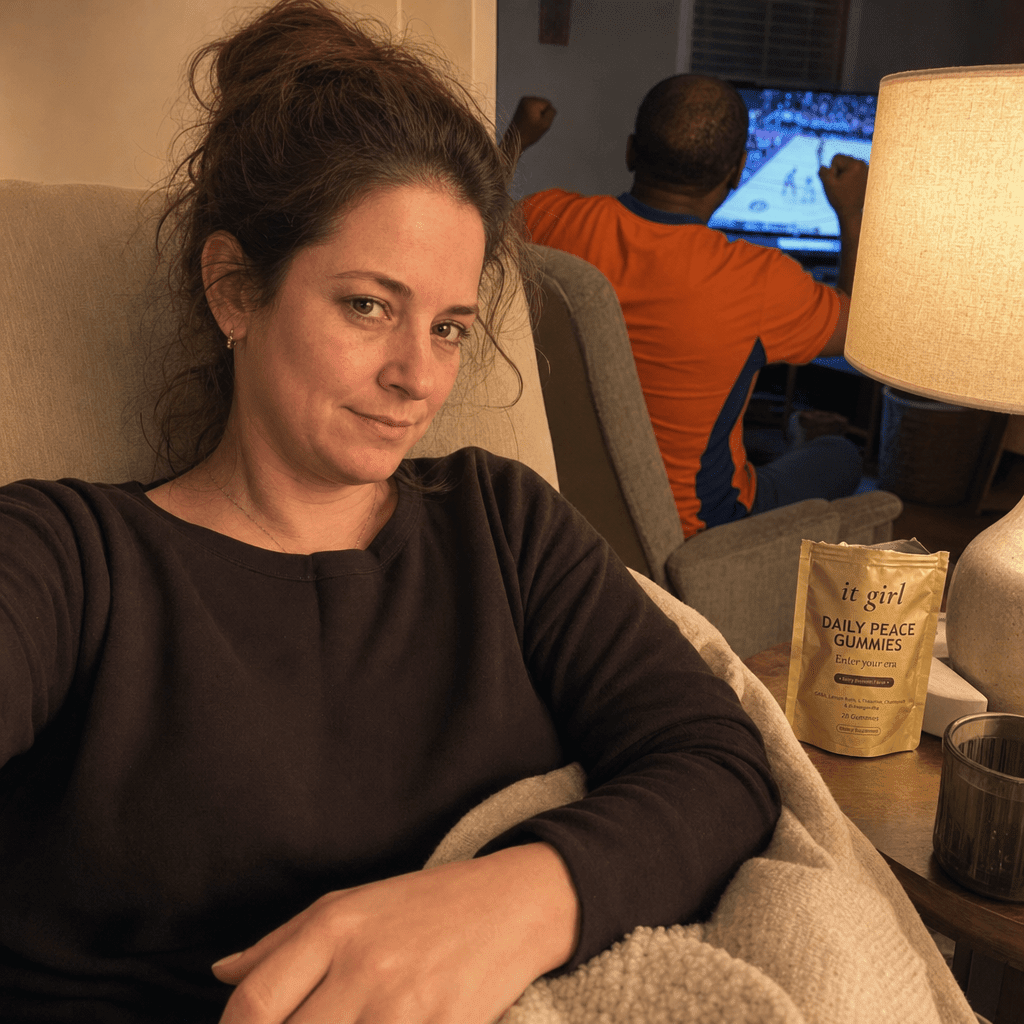 Calm mom on the couch during a blaring game, shoulders down and jaw unlocked, the real opened gold It Girl Daily Peace pouch resting on the side table beside her, her husband still animated behind her