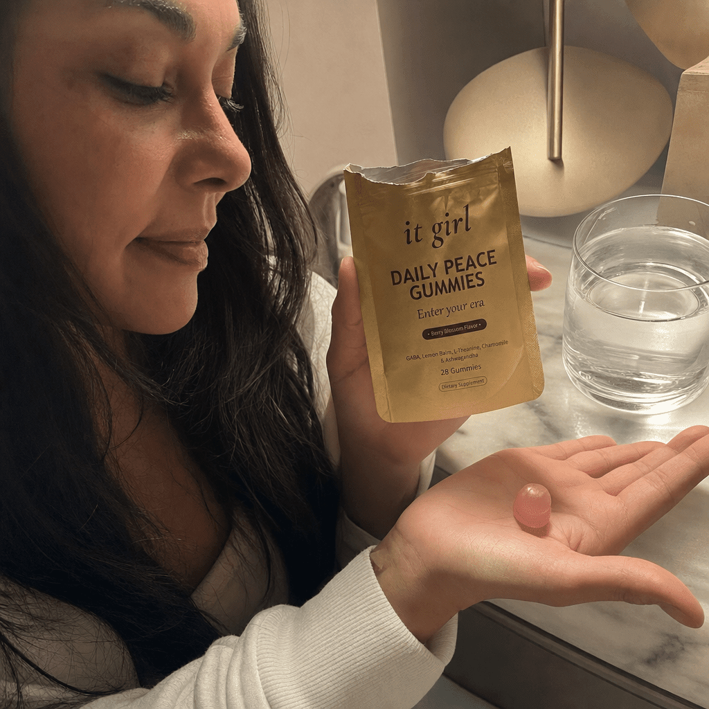 A woman's hand holding the opened matte-gold It Girl Daily Peace pouch with a single pink gummy in warm evening light beside a glass of water on a nightstand, a dollar-a-day evening ritual