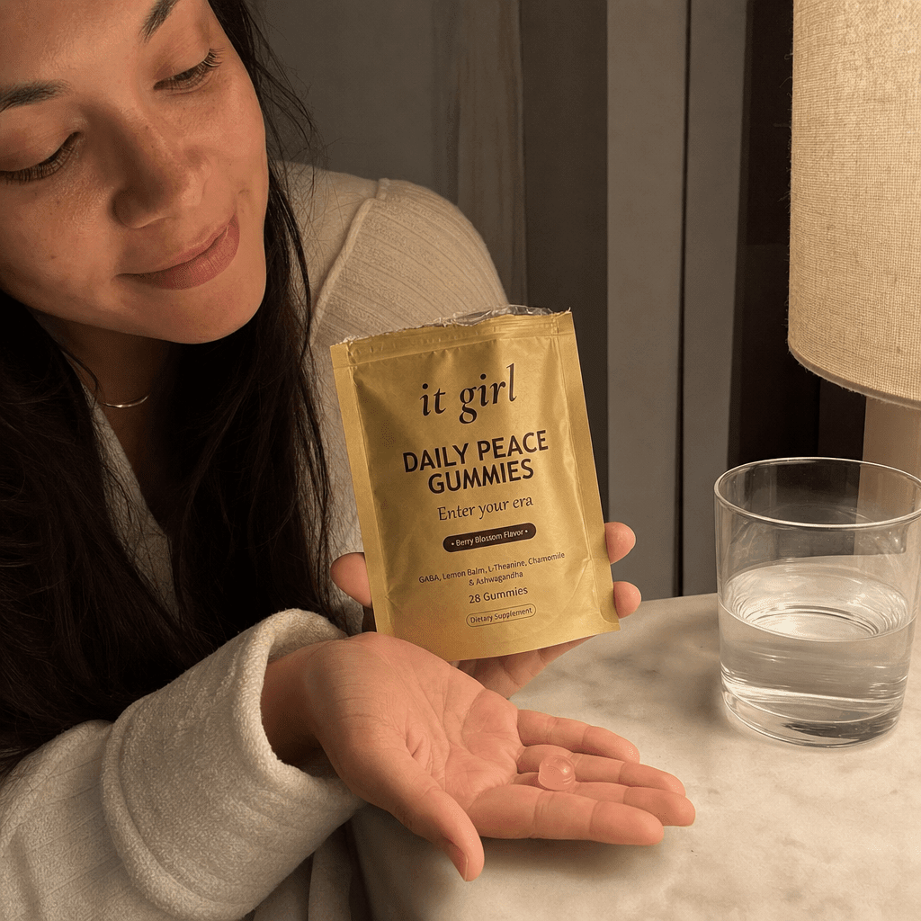 A woman's hand holding the opened matte-gold It Girl Daily Peace pouch with a single pink gummy in warm evening light beside a glass of water on a nightstand, a dollar-a-day evening ritual