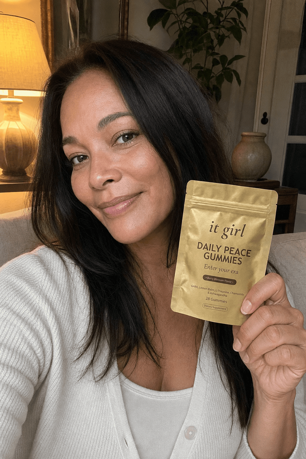 A calm, present woman in her forties in warm early-evening lamplight holding the real gold It Girl Daily Peace pouch with a relieved half-smile, no phone in sight
