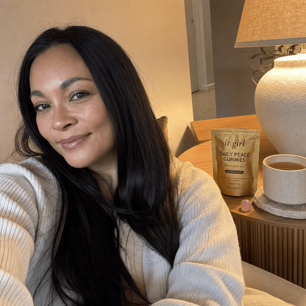 The same calm woman settled on her couch in warm early-evening lamplight with the gold It Girl Daily Peace pouch and a single gummy on the side table, calm not sedation, melatonin-free