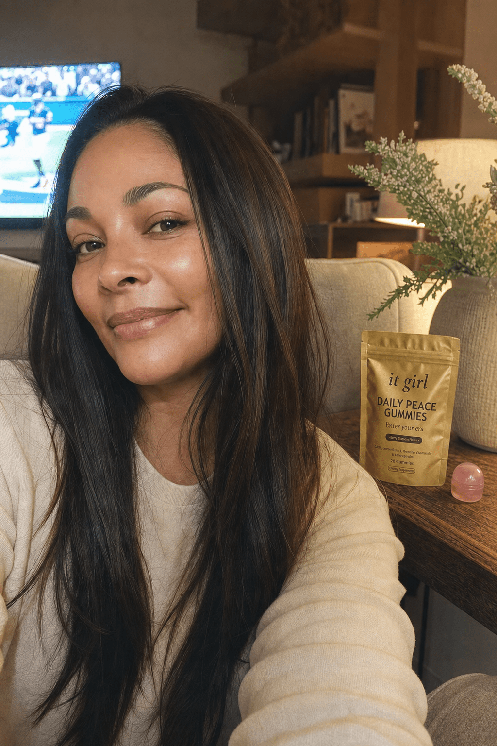 Cristina, a relaxed mom in her late thirties, present and at ease in her warm living room in the evening with the game softly on in the background, the real It Girl Daily Peace pouch on the side table