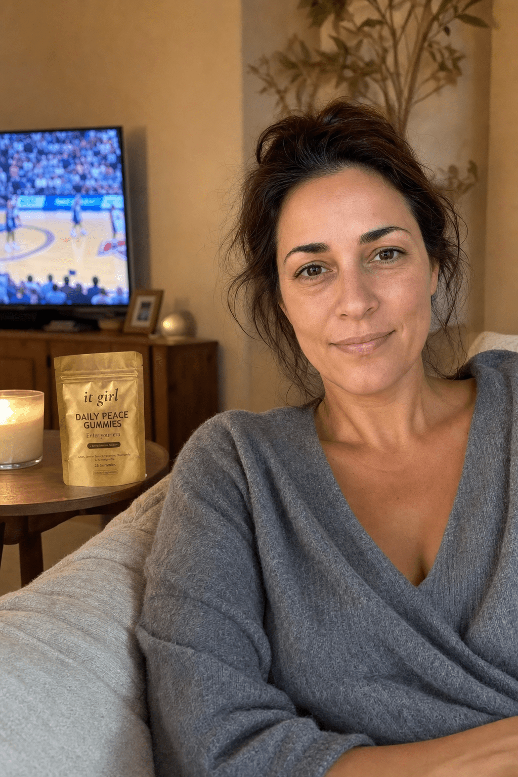 Warm candid evening photo of a put-together working mom about thirty-eight sitting easy on her own couch on game night, shoulders relaxed, the gold It Girl Daily Peace pouch on the side table beside her, the TV glowing soft and warm in the background
