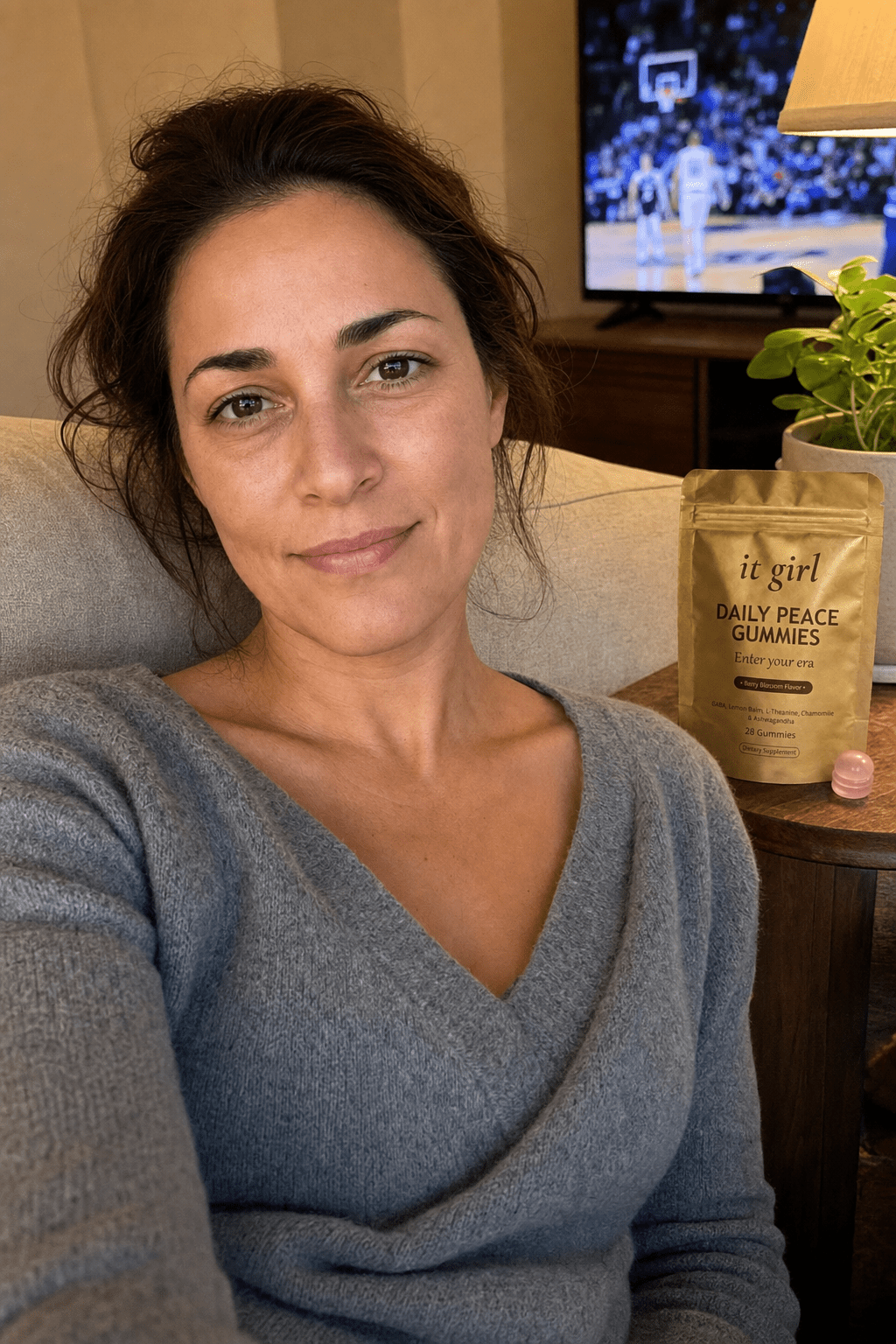 Warm candid evening photo of a put-together working mom about thirty-eight sitting easy on her own couch on game night, shoulders relaxed, the gold It Girl Daily Peace pouch on the side table beside her, the TV glowing soft and warm in the background
