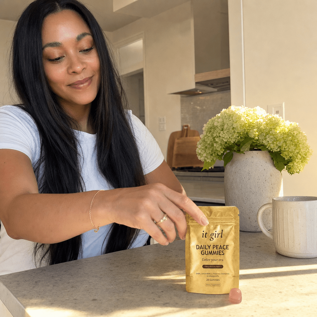 The real soft-gold It Girl Daily Peace pouch beside a single pink gummy and a glass of water on a warm cream surface, the calm signal handed over clean