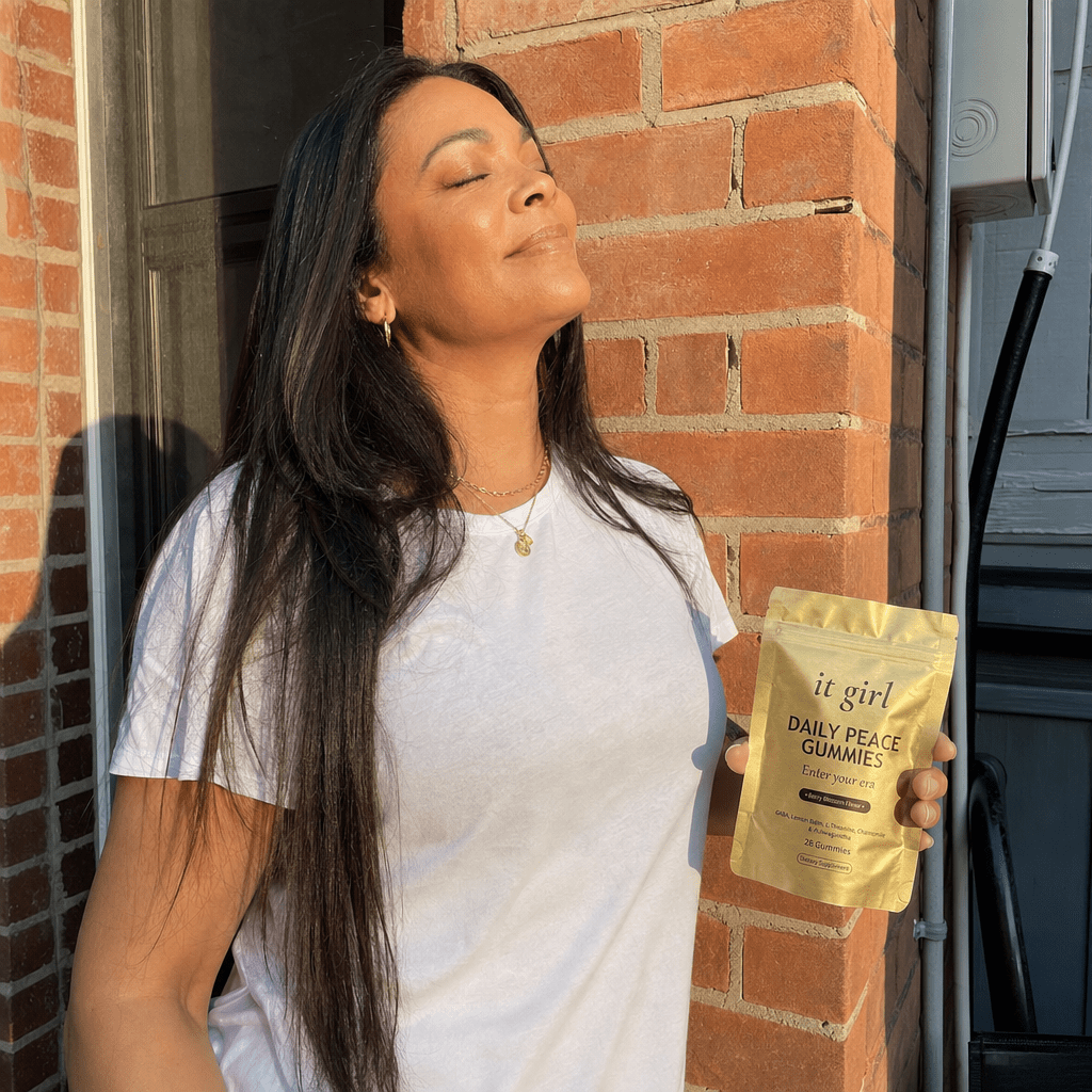 A woman in her forties stepping outside for her quiet five minutes, leaning against a sunlit doorway with her eyes closed and shoulders dropping as she exhales, the gold It Girl Daily Peace pouch in her relaxed hand
