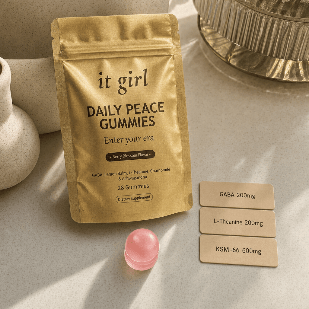 The matte gold It Girl Daily Peace pouch with a single pink gummy and three warm dose cards reading KSM-66 600mg, GABA 200mg, L-Theanine 200mg on a cream surface