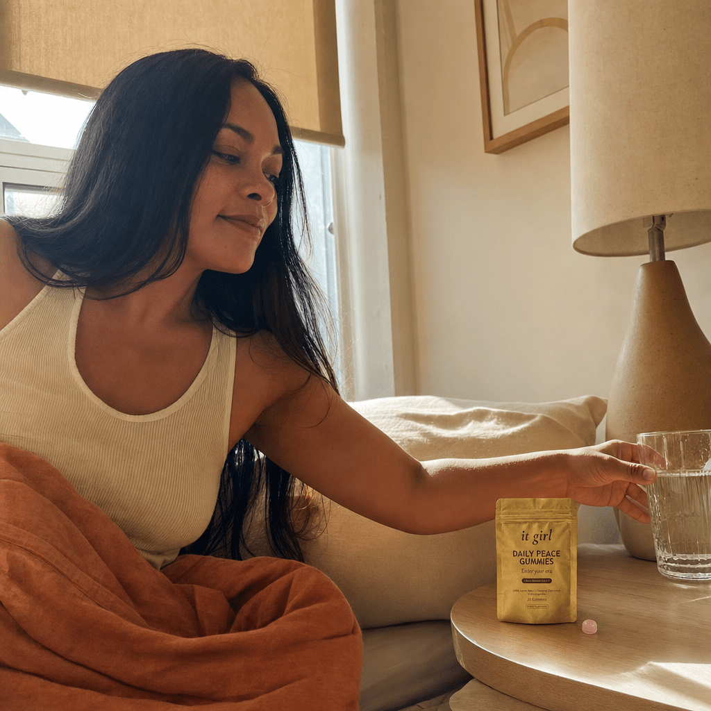 A woman waking up calm in soft morning light, reaching for a glass of water on the nightstand with the gold Daily Peace pouch nearby