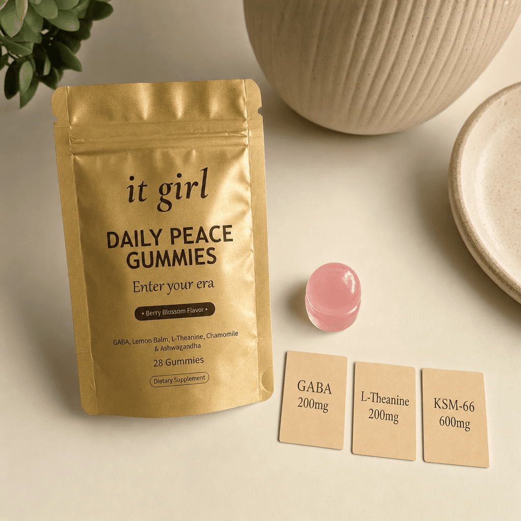 The matte gold It Girl Daily Peace pouch with a single pink gummy and three warm dose cards reading KSM-66 600mg, GABA 200mg, L-Theanine 200mg on a cream surface