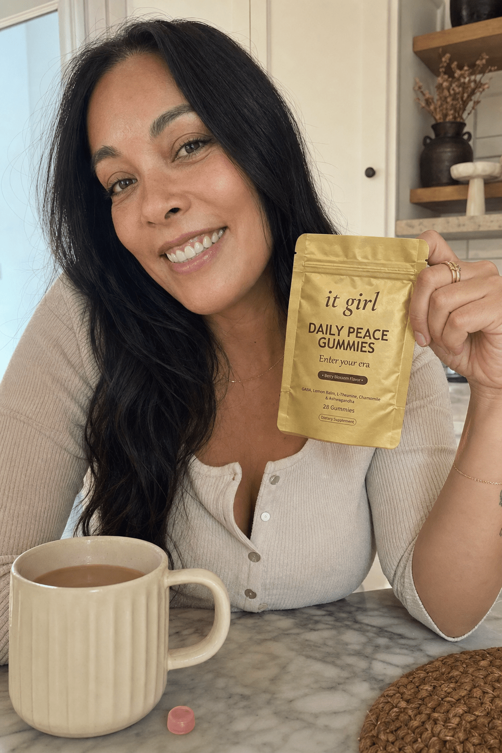 A calm woman in her forties at ease at her morning kitchen counter holding the gold It Girl Daily Peace pouch, settled and in command, coffee nearby