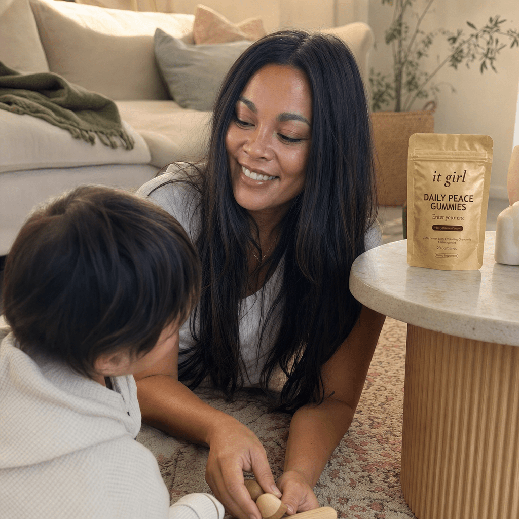 Mom in her forties down on the living-room floor fully present with her young child, relaxed and unhurried, the gold Daily Peace pouch resting on the side table nearby, soft warm light